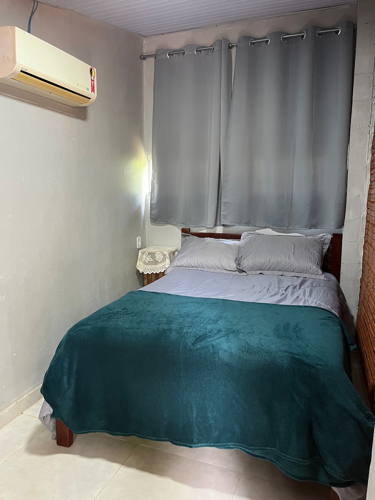 A compact sleeping area features a cozy bed draped with a soft dark teal blanket. Light gray pillows complement the bedding. A wall-mounted air conditioning unit is positioned above the bed, and the window is dressed with medium gray curtains, softly filtering natural light.