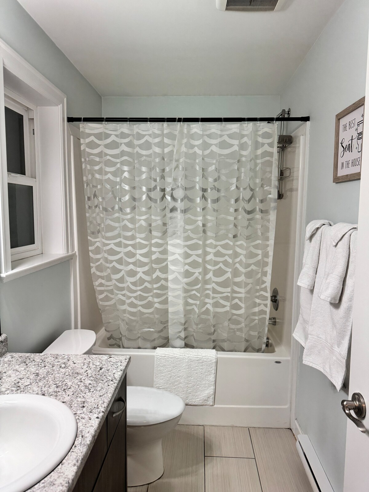 A clean bathroom is equipped with a shower and tub combination draped with a patterned shower curtain. A white toilet and a sink with a speckled countertop are situated nearby. Soft, white towels are neatly hung on a rack, and natural light is allowed in through a small window.