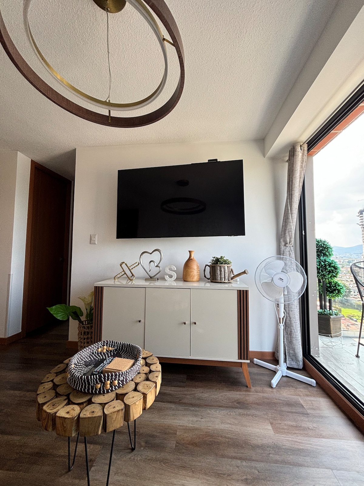A modern living area displays a wall-mounted television above a stylish cabinet with decorative items. A round wooden coffee table, featuring a textured centerpiece, complements the space. Natural light enters through the large glass doors, leading to a balcony with a view.