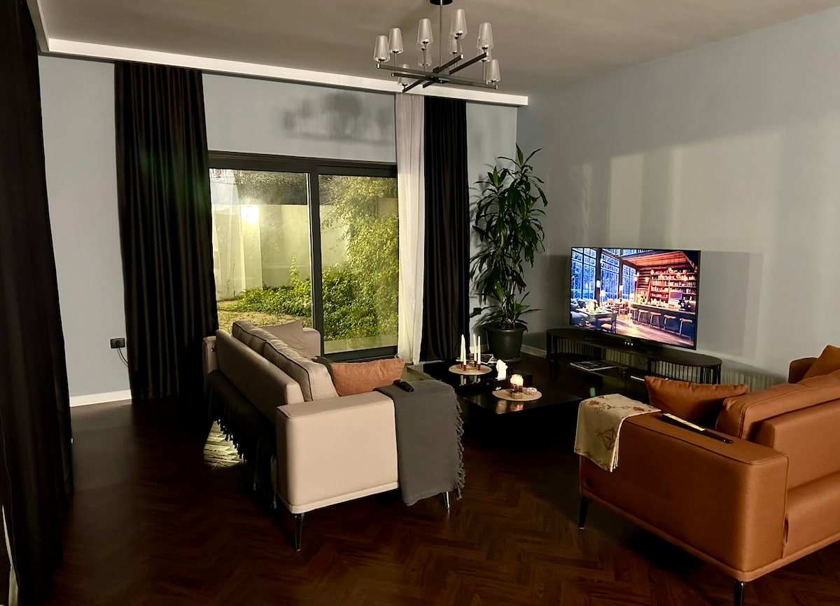 A stylish living area features a large window with soft lighting illuminating the space. Comfortable sofas are arranged with a coffee table displaying refreshments. A plant adds a touch of greenery, while a television is positioned across from the seating, enhancing the inviting atmosphere.