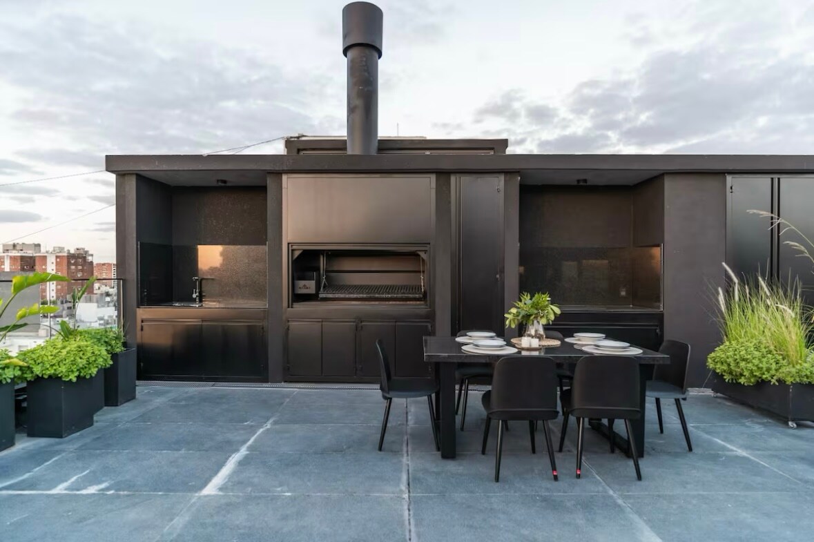 A modern rooftop terrace features a black grill and a dining area with a large table surrounded by chairs. Planters with greenery are placed along the edges, providing a touch of nature against the sleek design. The skyline is visible in the background.