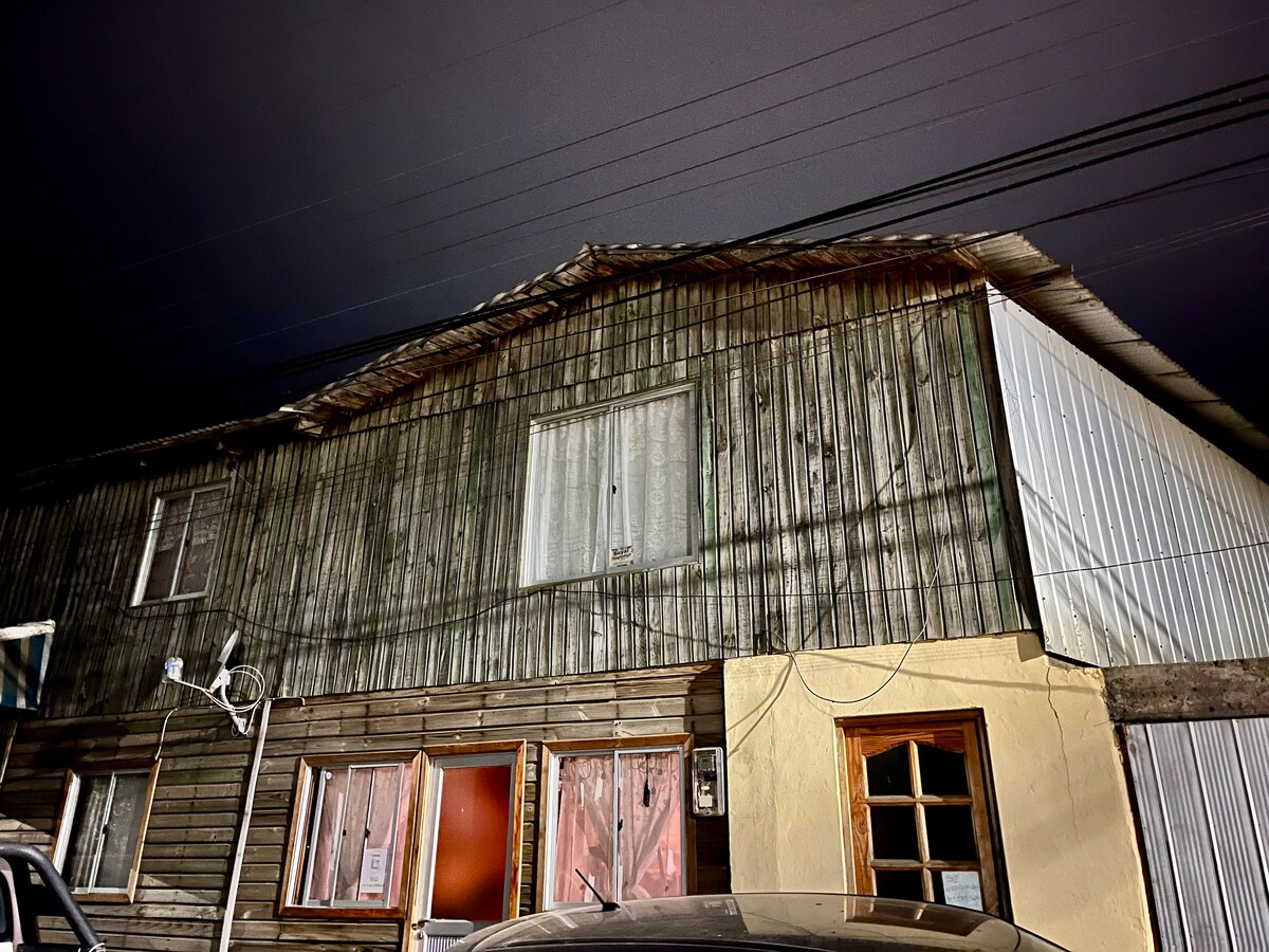 The exterior of a two-story building is captured at night, exhibiting a mix of wooden and metal siding. A large, light-colored window contrasts with the darker, textured walls. The entrance showcases a wooden door, and soft light glows through the curtains of the lower windows.