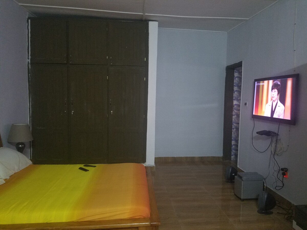 A spacious bedroom features a wooden bed covered with a bright orange and yellow gradient blanket. A flat-screen television is mounted on the wall opposite the bed. A lamp and a storage cabinet with closed doors are positioned near the bed, contributing to the room's simple decor.