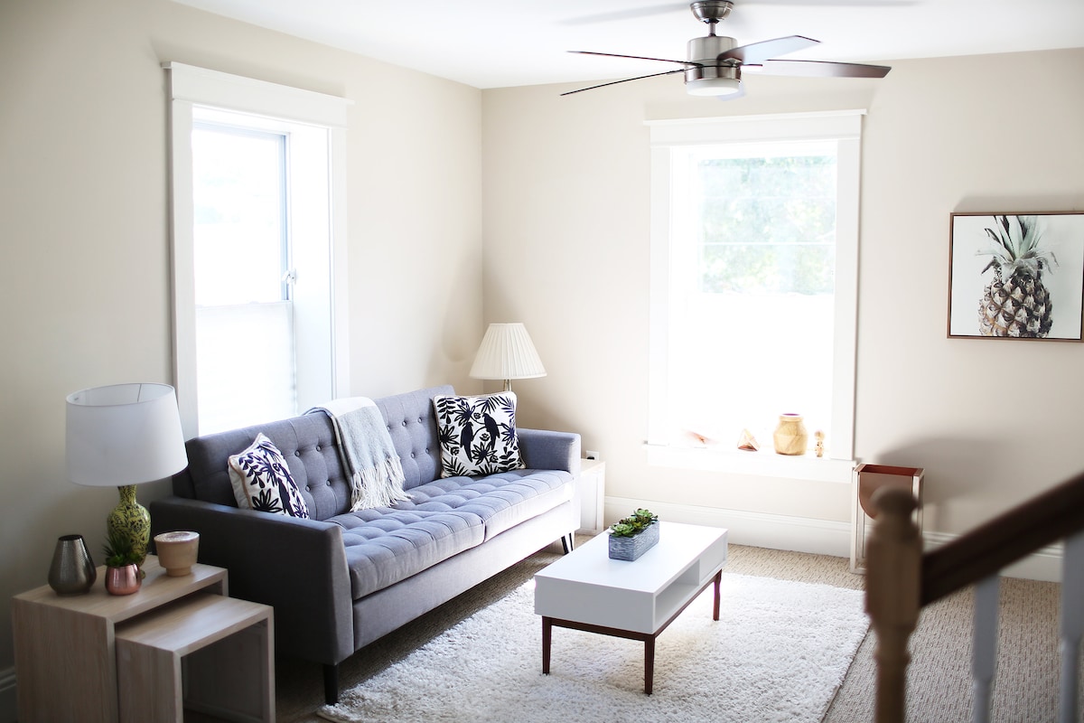 A light-filled living area features a gray tufted sofa adorned with decorative pillows. A white coffee table sits on a textured rug, with a small plant adding a touch of greenery. Two windows allow natural light to brighten the space, complemented by a modern ceiling fan.