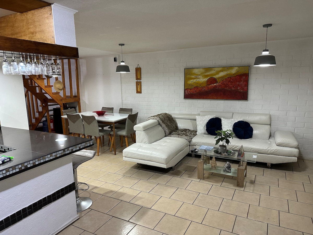 A comfortable living area features a white sectional sofa paired with blue accent pillows. An elegant coffee table sits atop tiled flooring, with a small plant on display. A dining table for six is situated nearby, complemented by art on the wall and modern pendant lighting.
