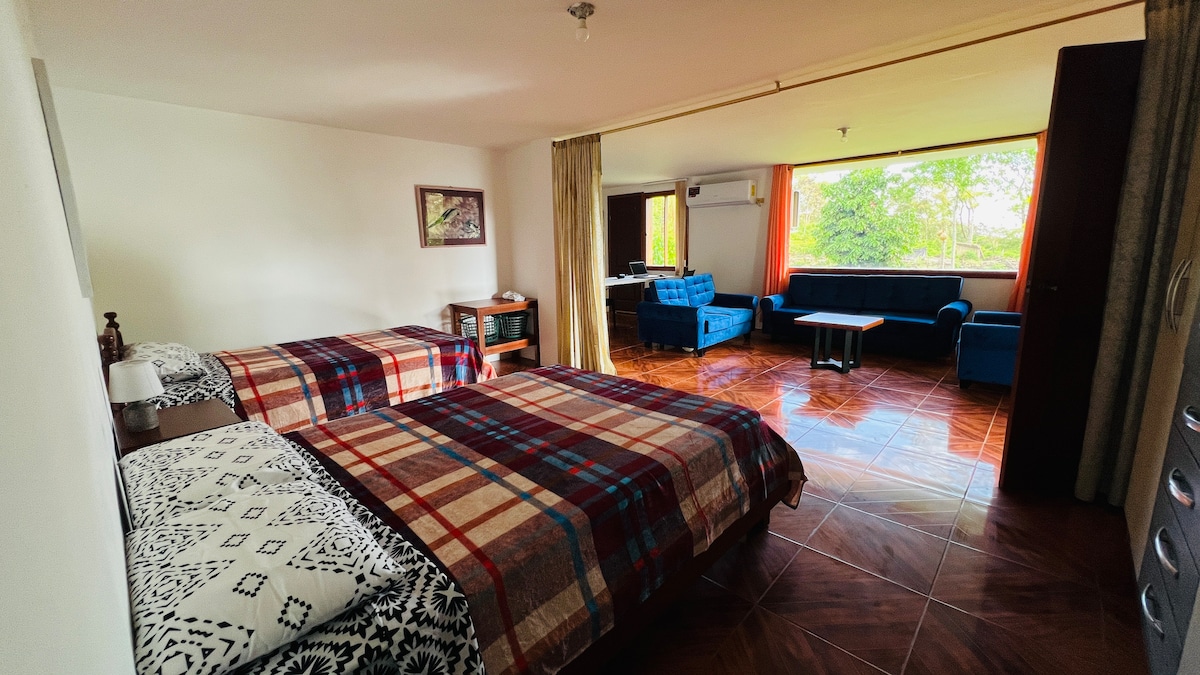 A spacious room is seen featuring two beds dressed in coordinated plaid and patterned bed covers. A lounge area with blue seating is visible in the background, alongside a large window offering views of the surrounding greenery. Natural light fills the room.