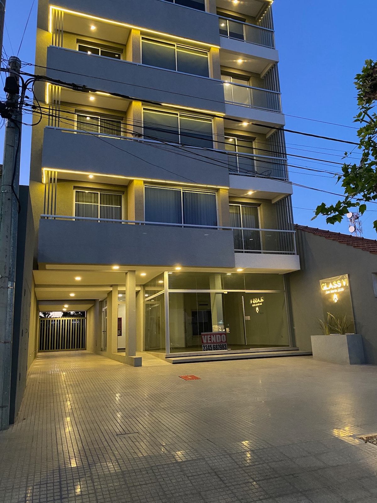 A modern four-story building is showcased in the image, featuring large glass windows and a well-lit entrance. The exterior is highlighted by soft, warm lighting, and the tiled pathway leads to a glass door, creating an inviting approach.