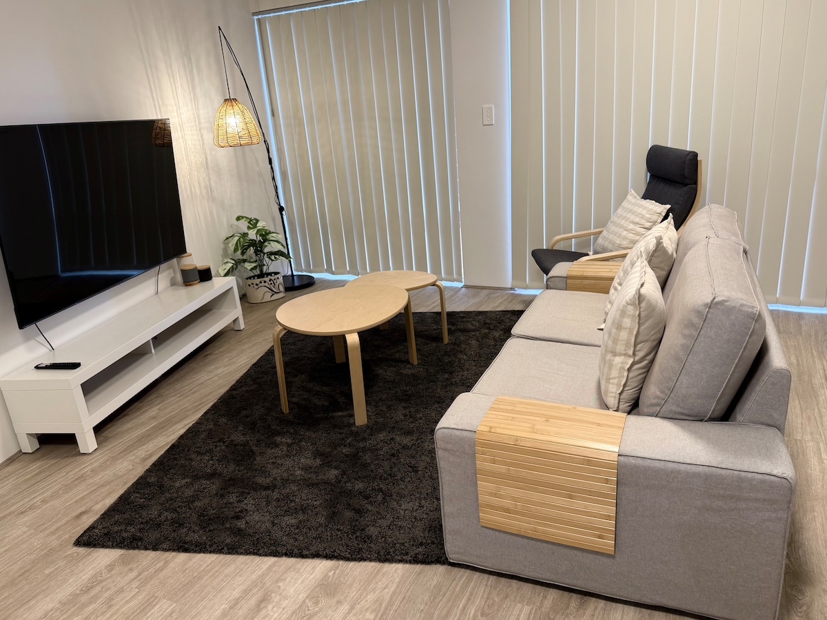 A spacious living area features a comfortable gray sofa and a single armchair, complemented by a round wooden coffee table. A large flat-screen TV is mounted on the wall. Natural light filters through vertical blinds, illuminating a nearby indoor plant.