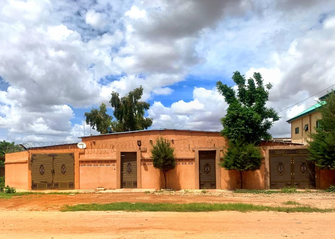 A single-story building with a textured ochre exterior features multiple decorative doors, each exhibiting intricate designs. Lush green trees flank the structure, and the sky is adorned with wispy clouds, creating a serene backdrop for this family-friendly space.
