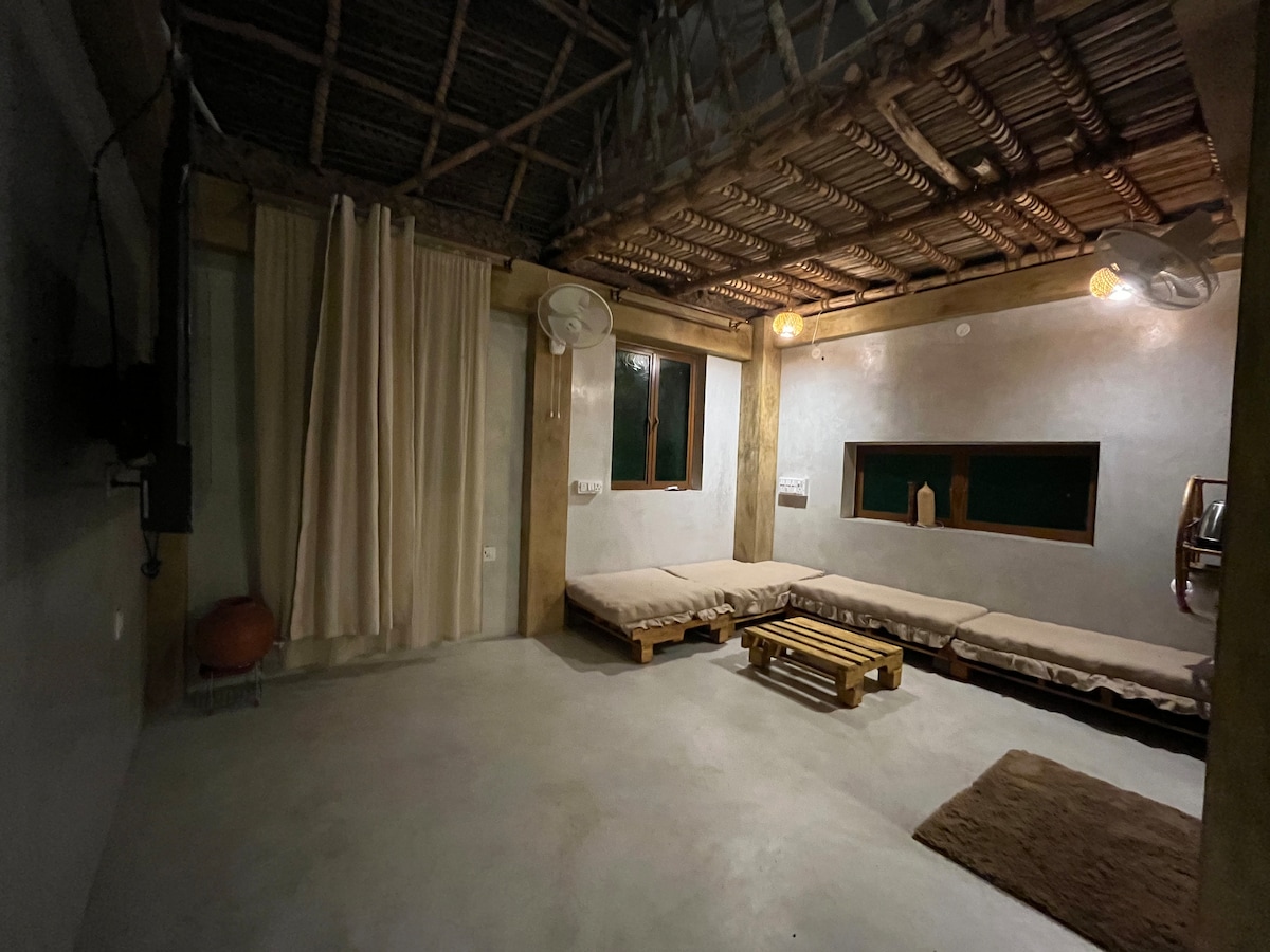 A spacious living area is showcased with a flat-screen TV mounted on the wall. Two wooden seating platforms are arranged, complemented by a wooden coffee table. Light filters through a large window, and soft curtains frame the space, creating an inviting atmosphere.