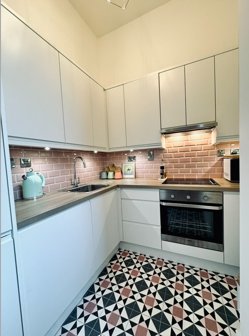 A modern kitchen is presented with sleek white cabinetry and a wooden countertop. A pink tiled backsplash complements the space. The kitchen includes a stainless steel oven and sink. Decorative items and a teal kettle are arranged on the countertop, with patterned flooring adding visual interest.