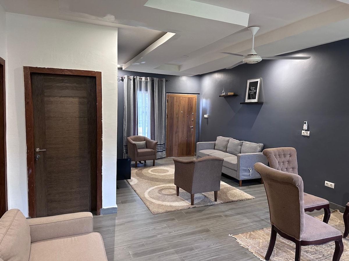A living area showcases a combination of cozy seating, including a pair of plush armchairs and a spacious gray sofa. A light-colored area rug sits on the floor, complementing the neutral tones of the walls. Natural light filters through the window, accentuated by elegant curtains.