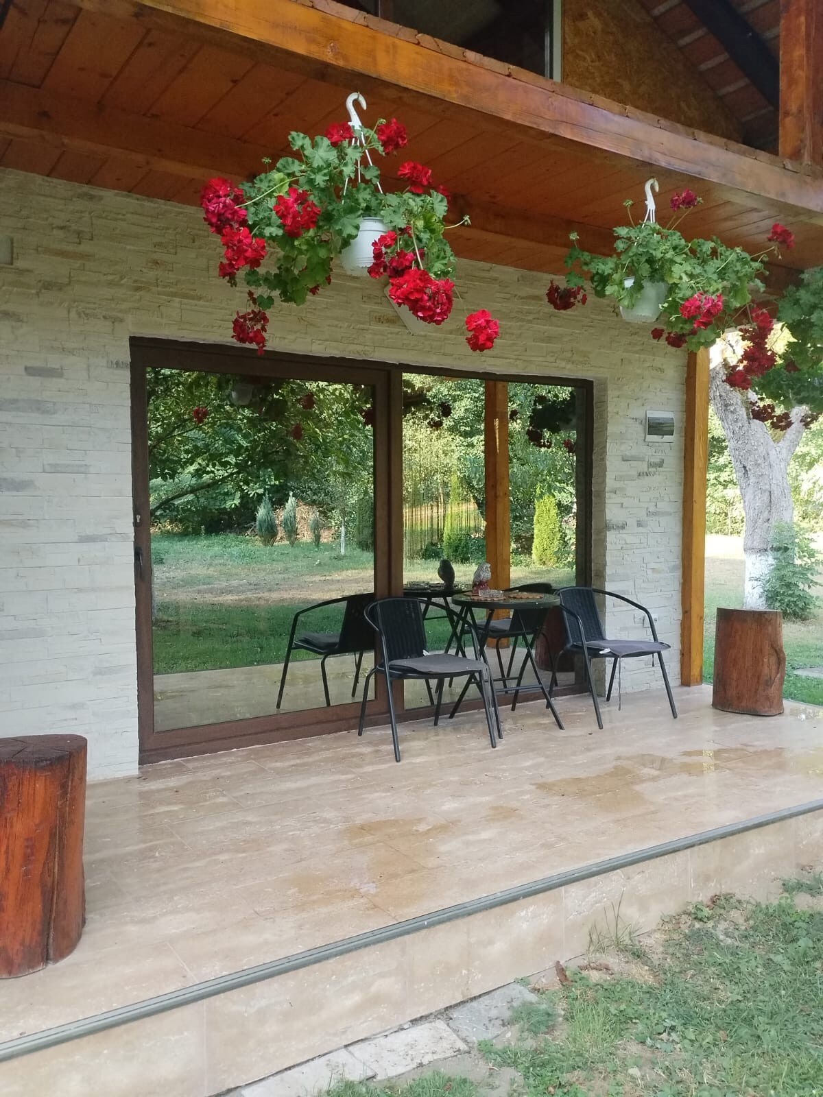 A spacious porch area is shown, featuring a set of four black chairs around a small table. Vibrant red flowers hang in decorative pots, complementing the natural surroundings of greenery visible beyond the glass doors.