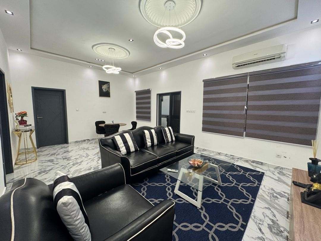 A modern living area features a sleek black leather sofa with decorative cushions, complemented by a glass coffee table. Two stylish ceiling fixtures provide illumination, with patterned window shades allowing natural light to filter in. Light-colored walls and a marbled floor enhance the spacious ambiance.