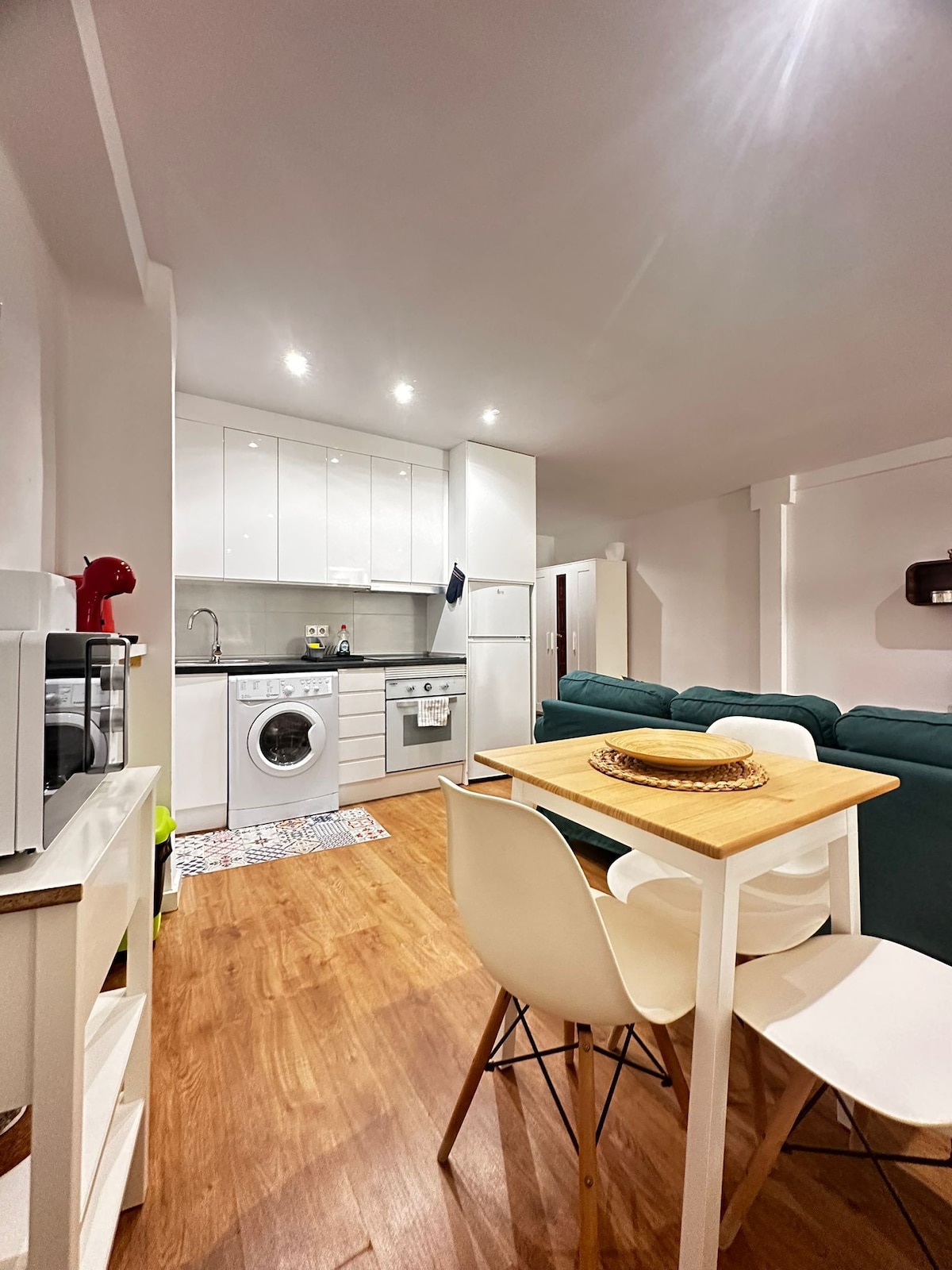 A modern kitchen space is presented, featuring sleek white cabinetry and essential appliances. A washer is located beside the stove. A small dining table is set for four, accompanied by white chairs. A green couch is visible in the living area, adding a touch of color to the space.