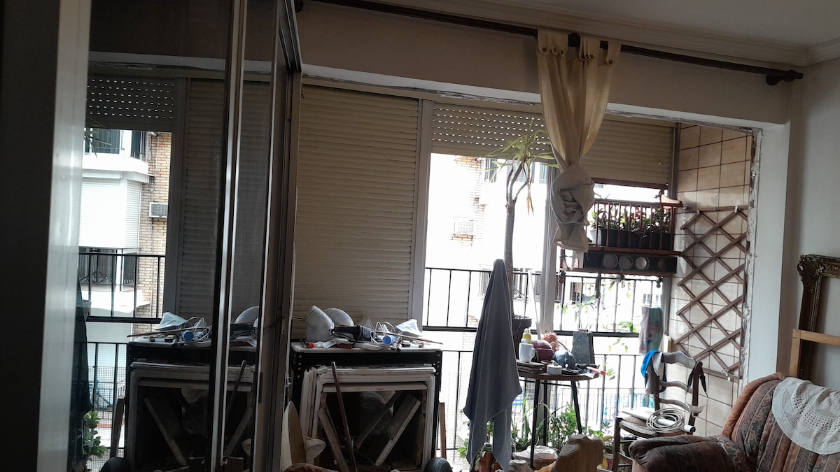 A view of a living area is presented, showcasing large windows that provide natural light. The space features a mix of furniture, including a chair and a small table, with items visible on shelves and a nearby balcony adorned with plants.