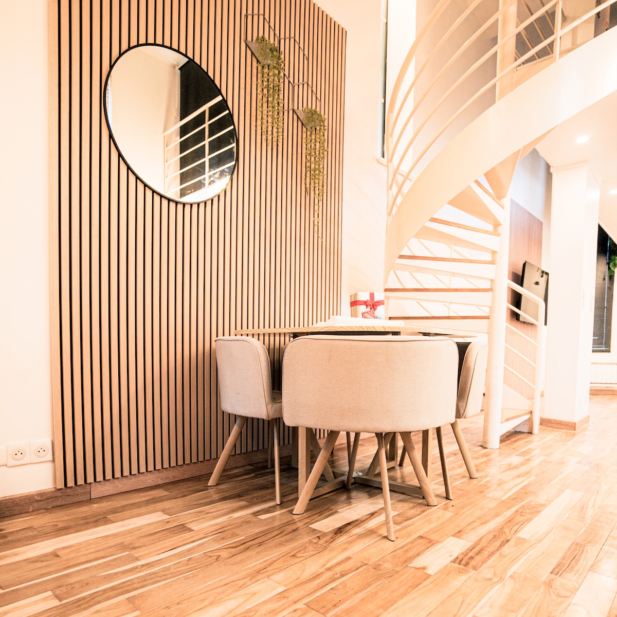 A dining area features a round wooden table surrounded by four light-colored upholstered chairs. A large round mirror is mounted on a slatted wooden wall, while a spiral staircase is visible in the background, enhancing the spaciousness of the room.
