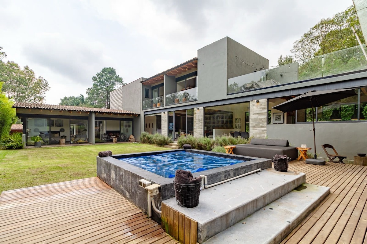 A modern home is set against a backdrop of green trees, featuring a spacious deck with a hot tub. The structure includes large glass windows and dark seating areas, creating a seamless connection with the surrounding landscape. The lawn is well-maintained, enhancing the overall serenity.