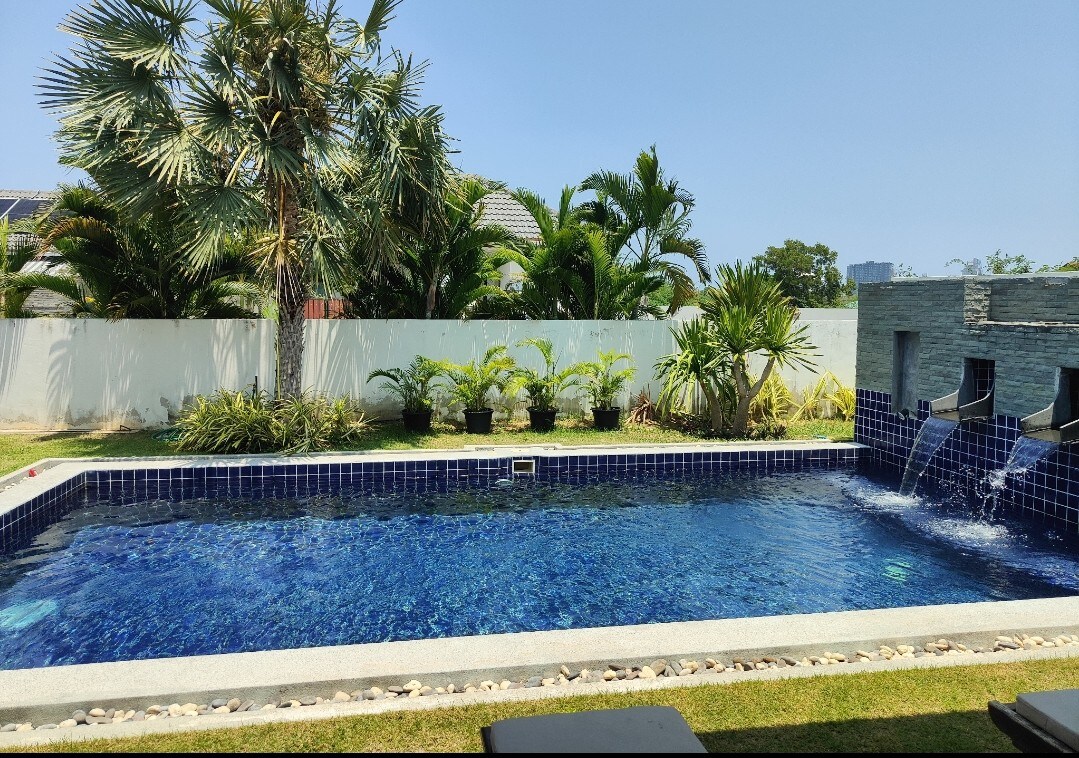 The inviting outdoor pool features a clear blue surface, surrounded by landscaped greenery. Water flows gracefully from wall-mounted spouts into the pool, set against a backdrop of palm trees and lush plants, providing a tranquil atmosphere.