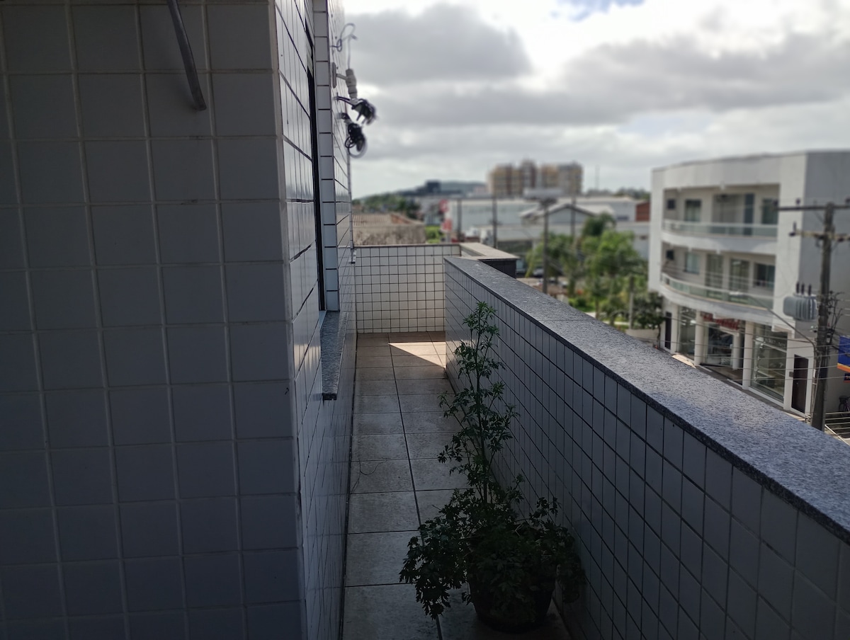 A balcony area is shown with tiled flooring and a low wall. A potted plant sits near the edge, and surrounding urban scenery, including buildings and streets, is visible in the background. The sky is partly cloudy, contributing to a bright atmosphere.