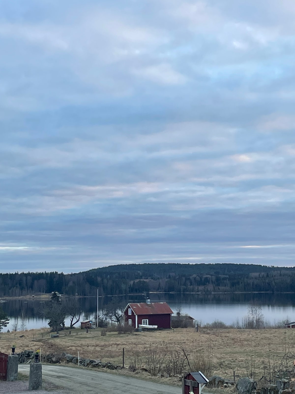 A scenic view captures a charming red house set against a backdrop of tranquil waters and rolling hills. The sky is filled with soft clouds, reflecting the serene environment surrounding the property, with trees lining the shore and greenery in the distance.