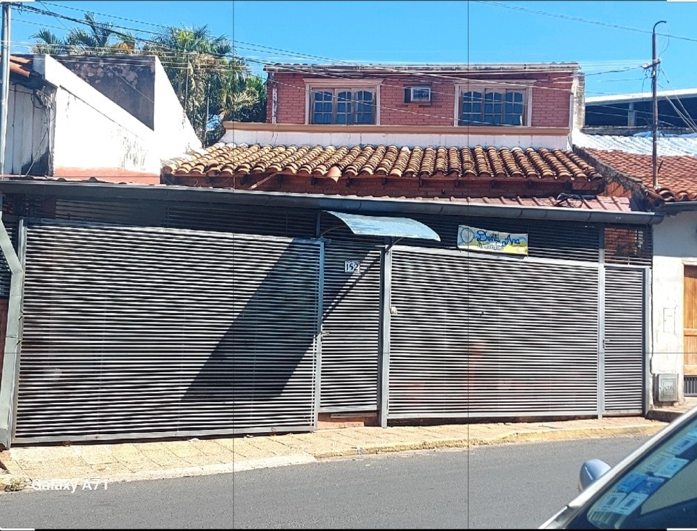 A residential building features a traditional facade, characterized by a terracotta roof and rolling metal shutters. A sign is visible above the entrance, indicating the presence of a business. Surrounding elements include nearby trees and a paved walkway.