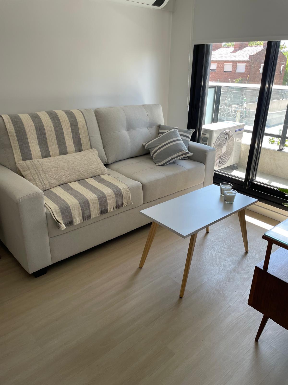 A light-filled seating area features a comfortable sofa with a striped throw, accompanied by a small coffee table. Large windows provide access to natural light and a view of the balcony, while an air conditioning unit is visible on the wall.