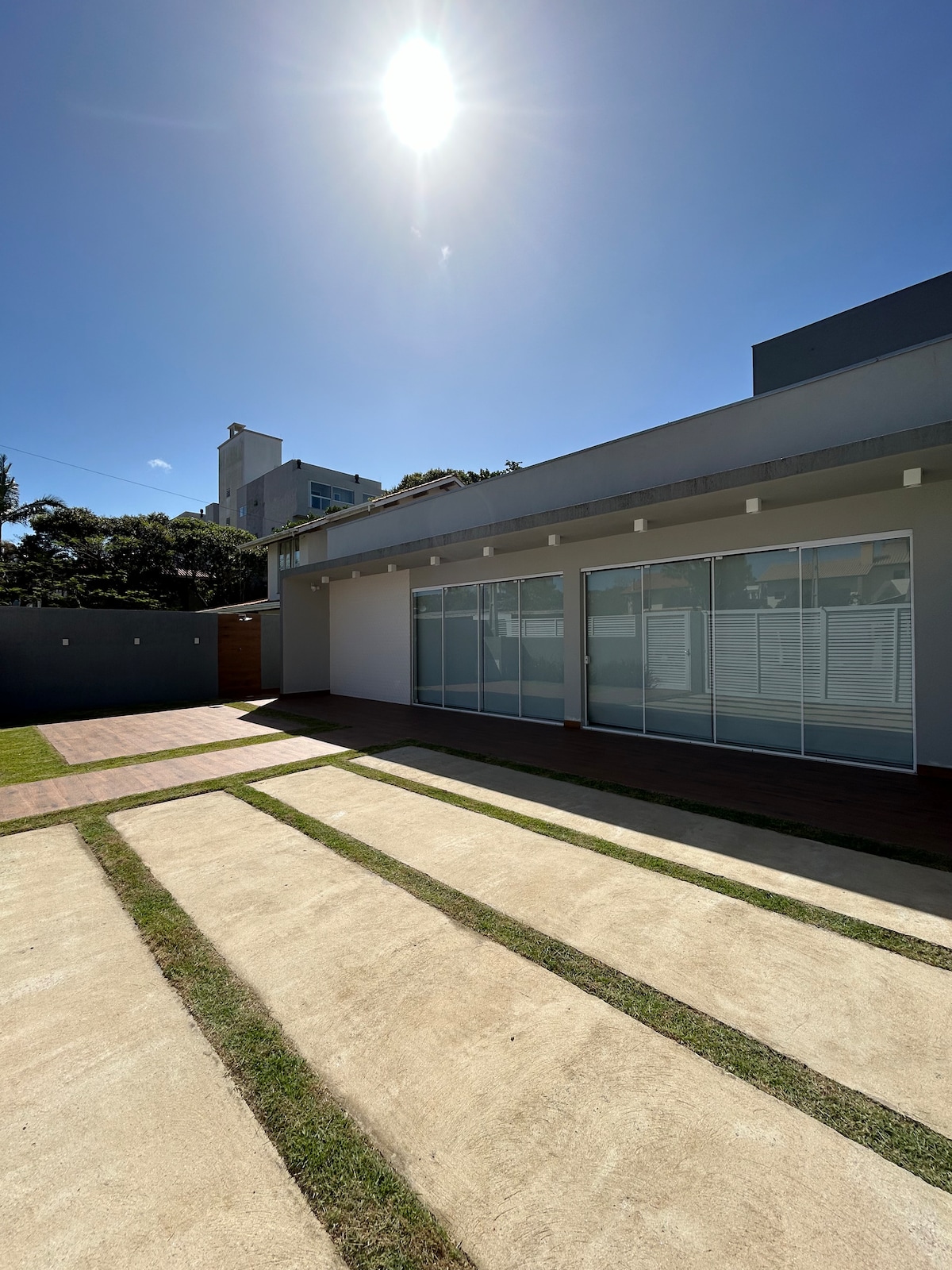 The spacious outdoor area is highlighted by a smooth pathway leading toward the house. Large glass doors allow natural light to flood the interior. The surrounding grass is neatly maintained, and the clear blue sky is visible overhead.