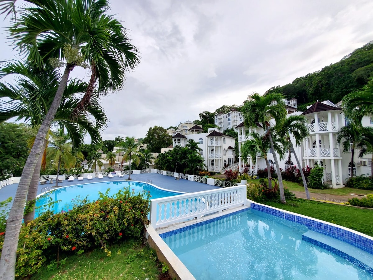Two swimming pools are visible, surrounded by lush greenery and tropical plants. The larger pool features a light blue hue, while the smaller one displays a deeper blue. Several lounge chairs are positioned near the pools, and a multi-story building is seen in the background.