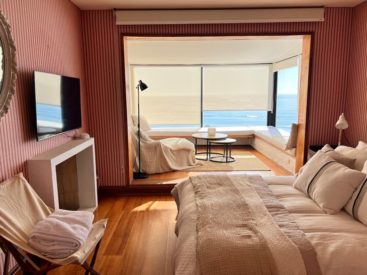 A cozy bedroom features a spacious bed with soft linens, complemented by warm wooden flooring. A large window offers a clear view of the ocean. A seating area with light-colored cushions is positioned next to the window, inviting relaxation and natural light.