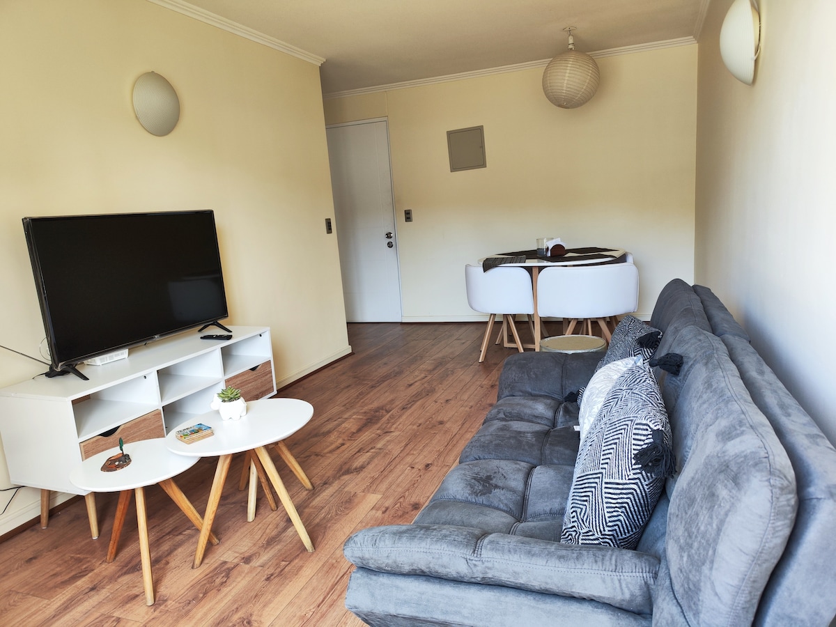 A living area features a gray sofa and a white coffee table with wooden legs. A television is positioned on a white media console. A dining area with a round table and white chairs is visible in the background, creating a balanced and functional space.