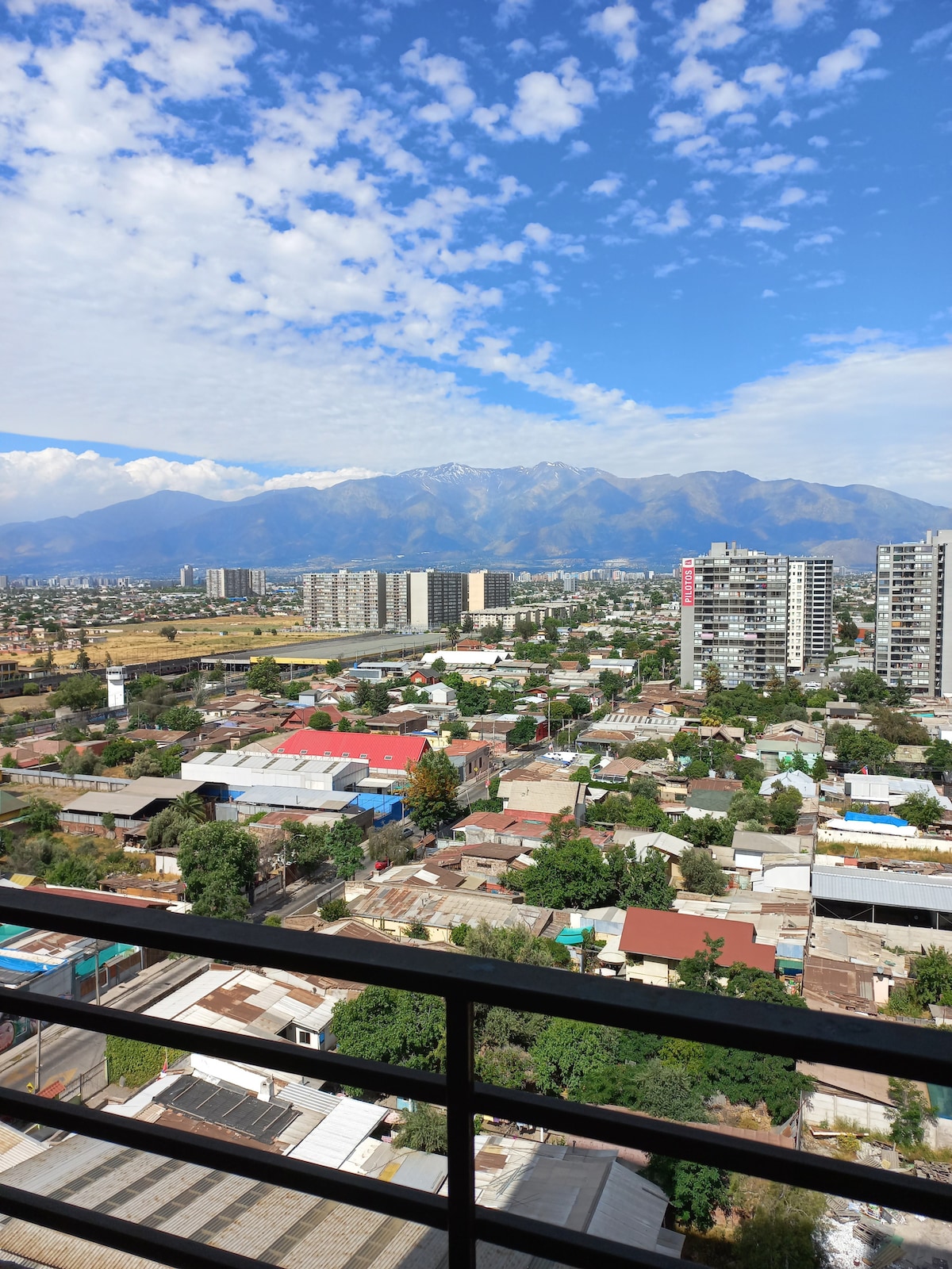 A panoramic view showcases a vibrant cityscape with a mix of residential buildings and greenery. Majestic mountains rise in the background under a blue sky dotted with clouds, offering a serene contrast to the urban landscape.