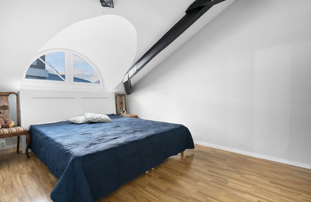 A room features a spacious bed dressed in a dark blue coverlet, accompanied by decorative pillows. Natural light enters through a large, arched window above the bed, highlighting the smooth, light hardwood floor and simple white walls.
