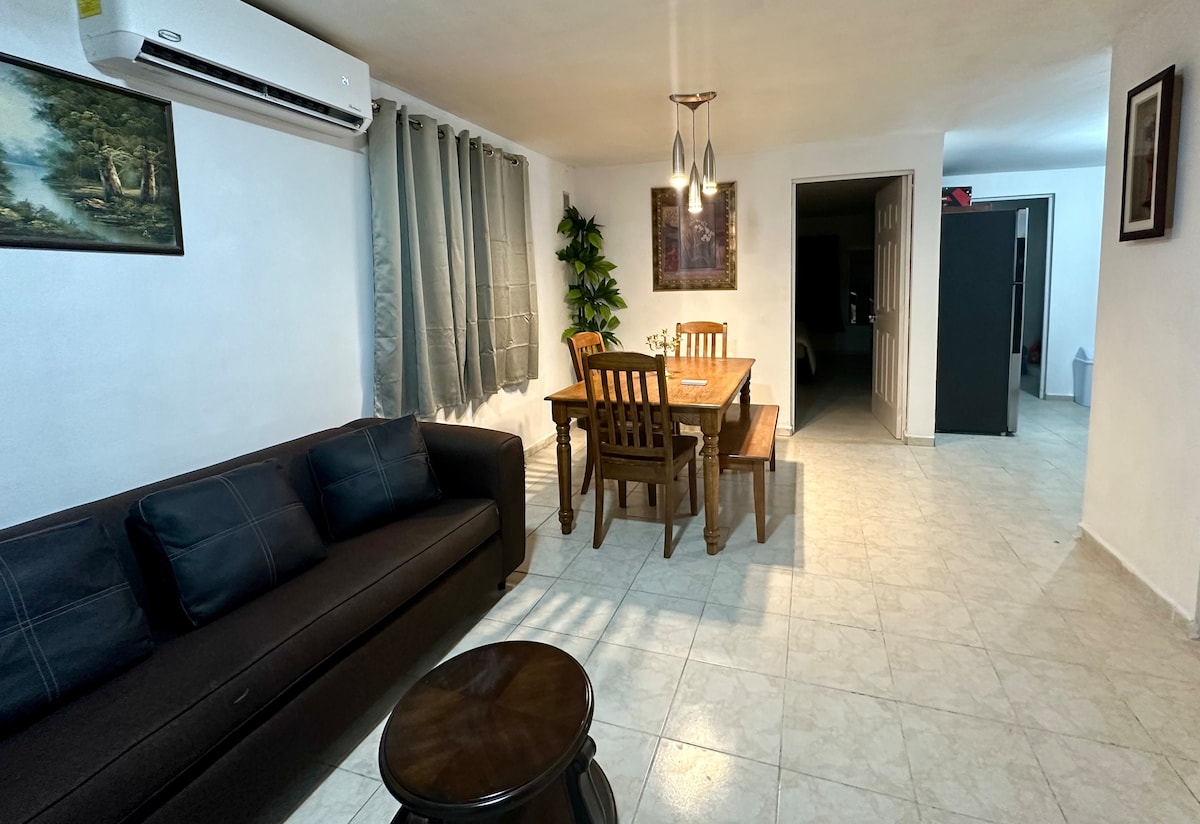 A comfortable living area features a dark sofa with subtle patterns, paired with a dining table and chairs. Natural light enters through window coverings, highlighting the tiled floor. Artwork and a decorative plant enhance the space, which is equipped with air conditioning.