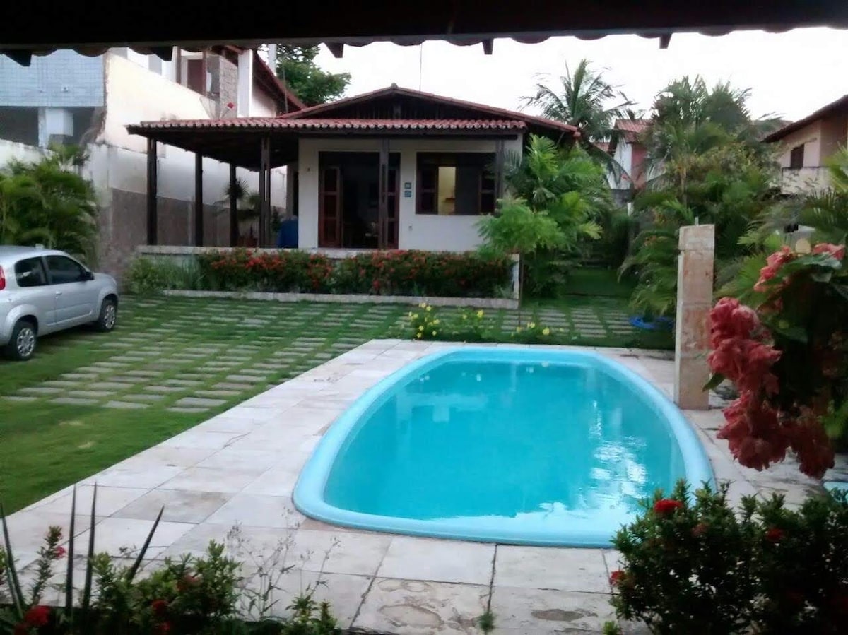 A landscaped outdoor area is highlighted by a clear swimming pool, surrounded by neatly arranged stone tiles and vibrant green grass. Lush tropical plants frame the space, while a covered seating area is visible in the background, providing shaded shelter.