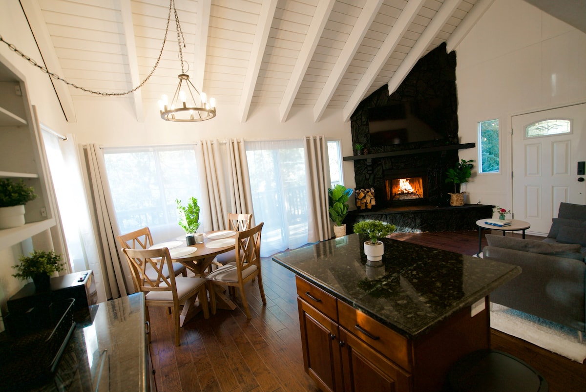 An inviting living space features a vaulted ceiling with exposed beams and large windows allowing natural light. A stone fireplace is centered on one wall, while a round dining table with four chairs and a cozy seating area are visible. Dark wood flooring enhances the warm ambiance.