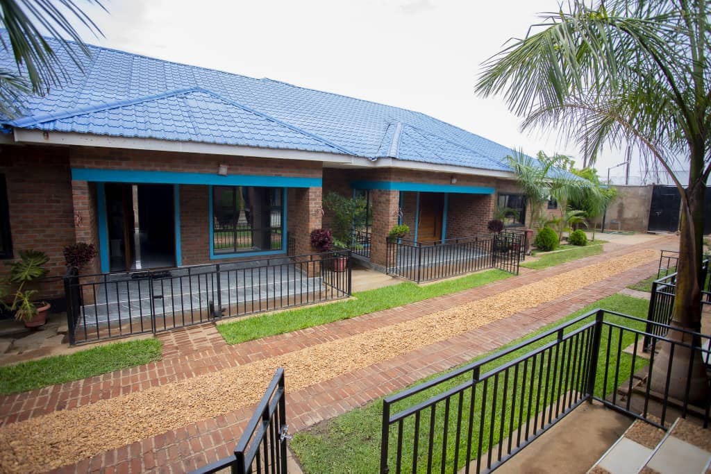 A well-maintained pathway lined with decorative plants leads to the entrance of multiple residential units. Each unit features a brick facade with blue accents and a tiled roof. Lush greenery complements the space, creating a serene environment.