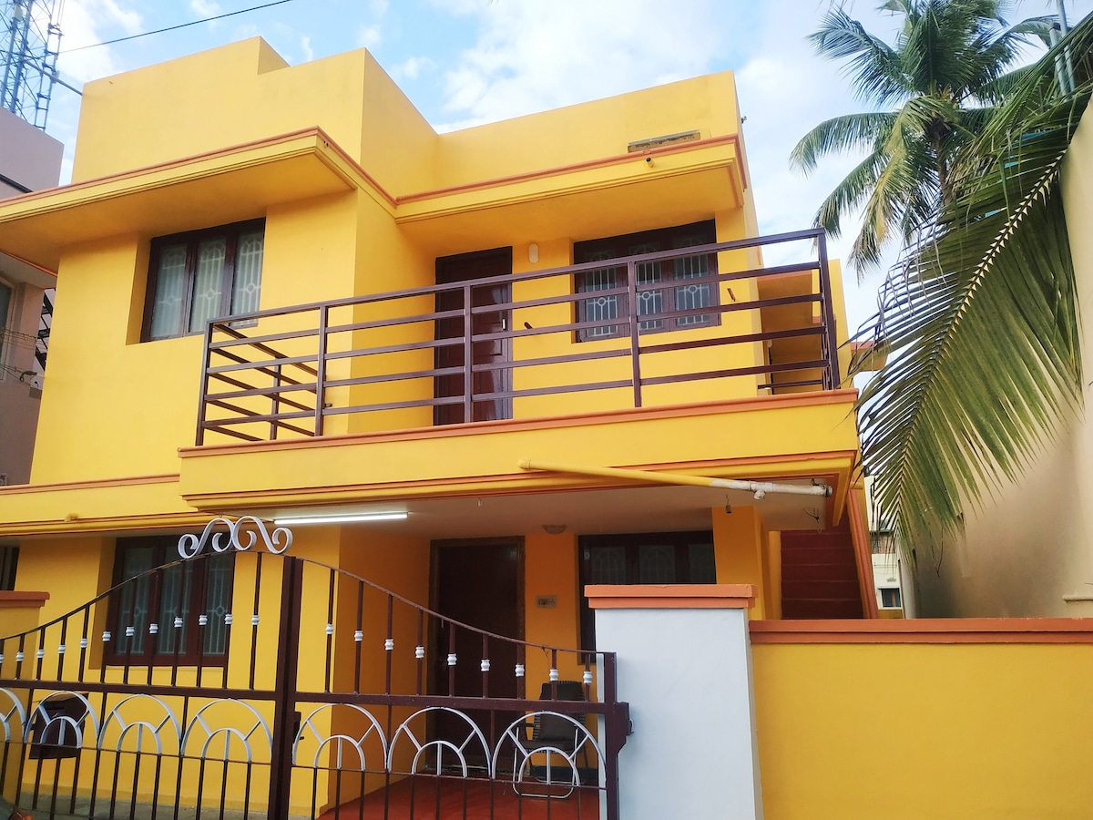 The exterior of a vibrant yellow two-story home is displayed, featuring a balcony and a sturdy railing. Lush green palm leaves are visible, enhancing the tropical vibe. A secure gate, leading to the entrance, complements the welcoming appearance of the property.