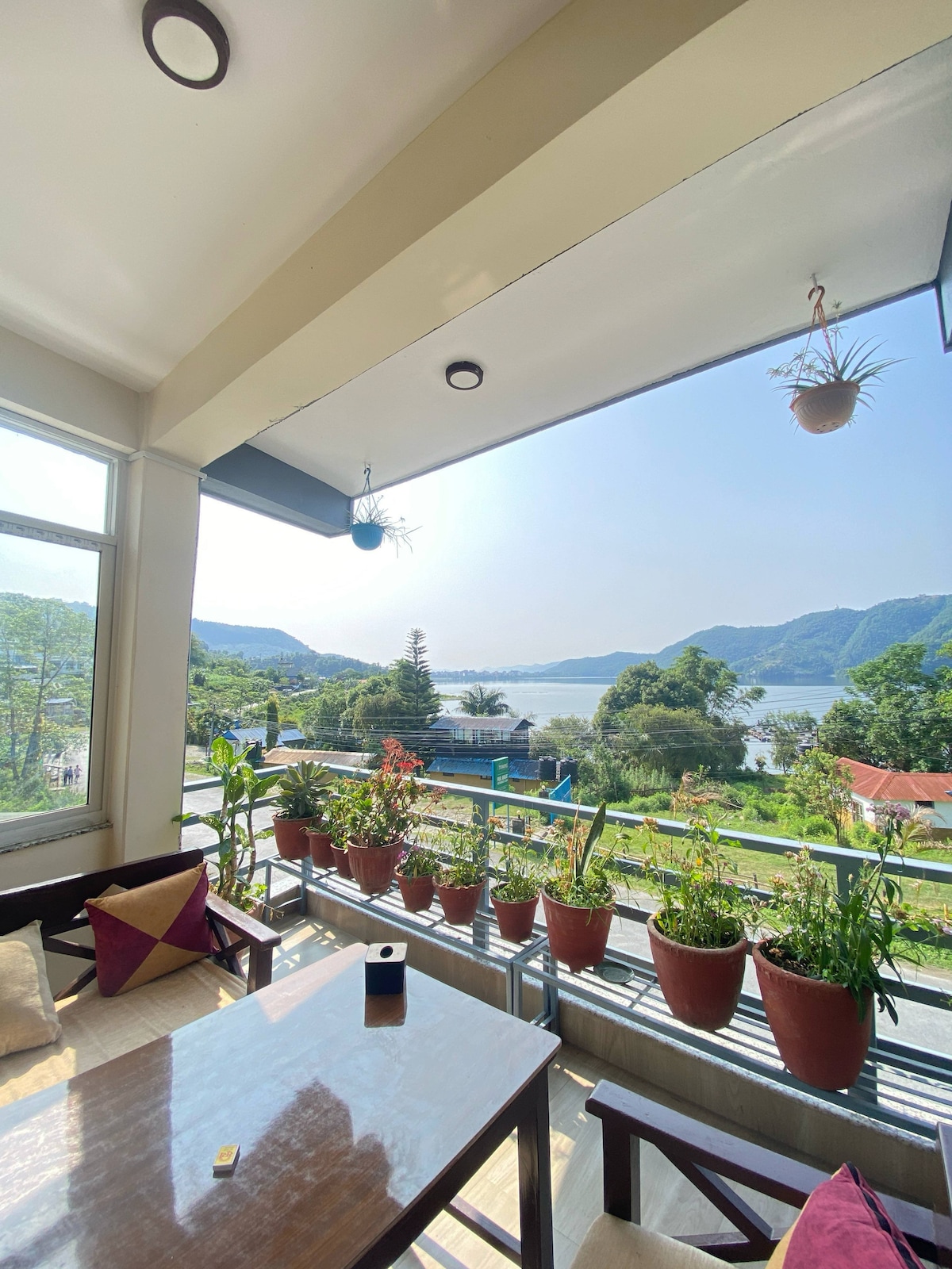 A spacious balcony is featured, adorned with a variety of potted plants along the railing. A wooden table and chairs provide a comfortable seating area. Scenic views of Fewa Lake and surrounding green hills are visible in the background under a clear blue sky.