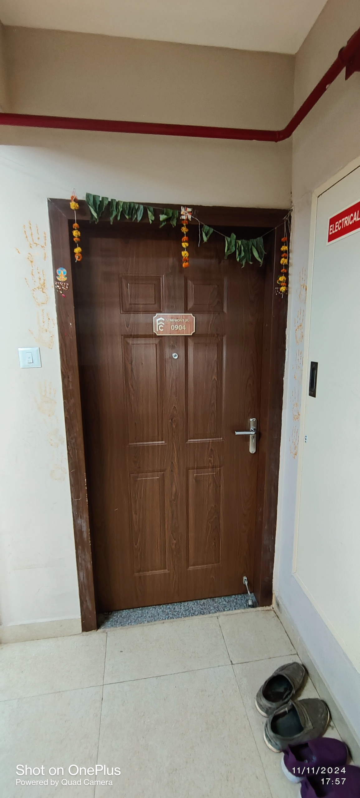 A wooden door features a small sign displaying the room number, adorned with decorative garlands made of flowers and greenery. A doormat is positioned at the entrance, and a pair of shoes rests nearby on the tiled floor.