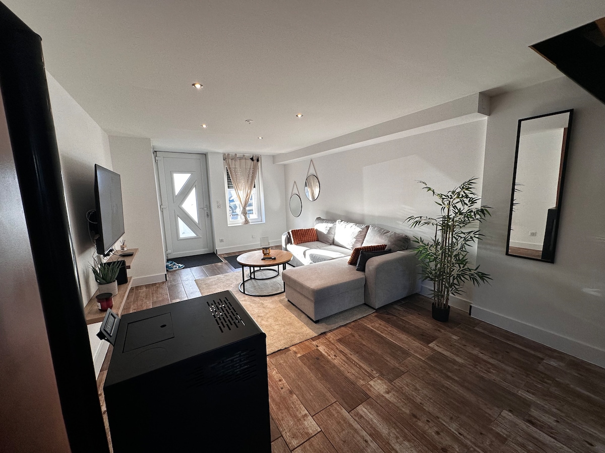 An open living area features a light-colored sofa arranged near a coffee table on a textured rug. A large mirror and decorative plants enhance the space. Natural light enters through the glass door, which leads to the outside, complemented by warm overhead lighting.