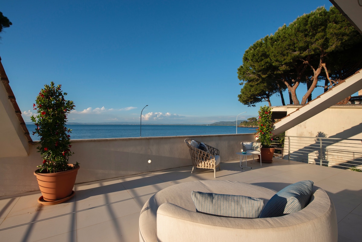 A spacious terrace features a circular lounge area adorned with cushions, providing a perfect spot for relaxation. Potted greenery adds a touch of nature, while a clear view of the sea extends beyond the railing, creating an inviting outdoor space bathed in natural light.