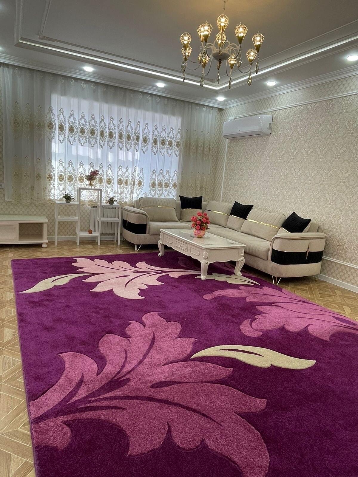 A well-lit living area features a large, plush purple area rug with floral patterns. A modern sofa with black and beige accents is arranged around a low coffee table, which holds a small vase of flowers. Sheer curtains dress the window, allowing natural light to enhance the space.
