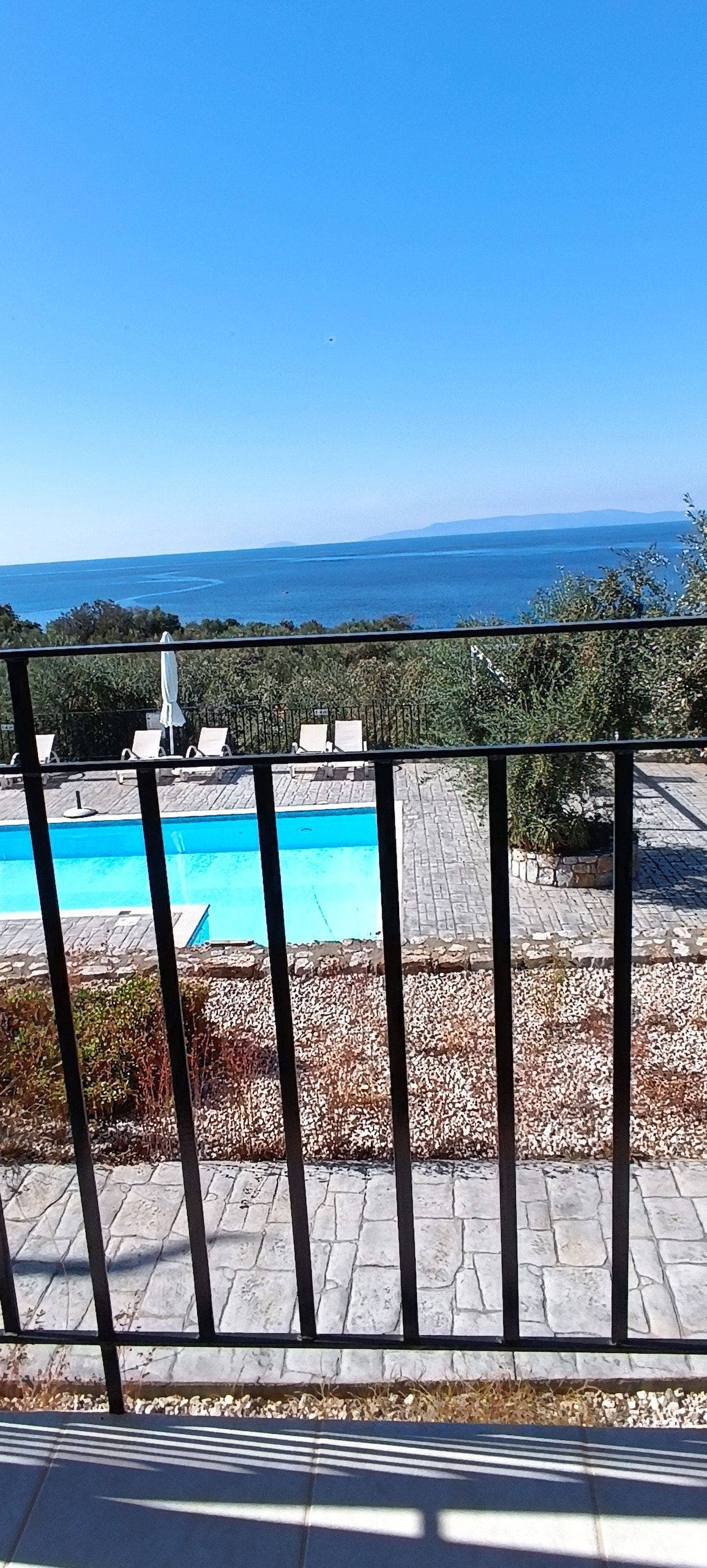A view of a serene outdoor area featuring a clear swimming pool surrounded by neatly arranged stones. In the distance, a calm sea extends towards the horizon, framed by greenery and sunny blue skies.