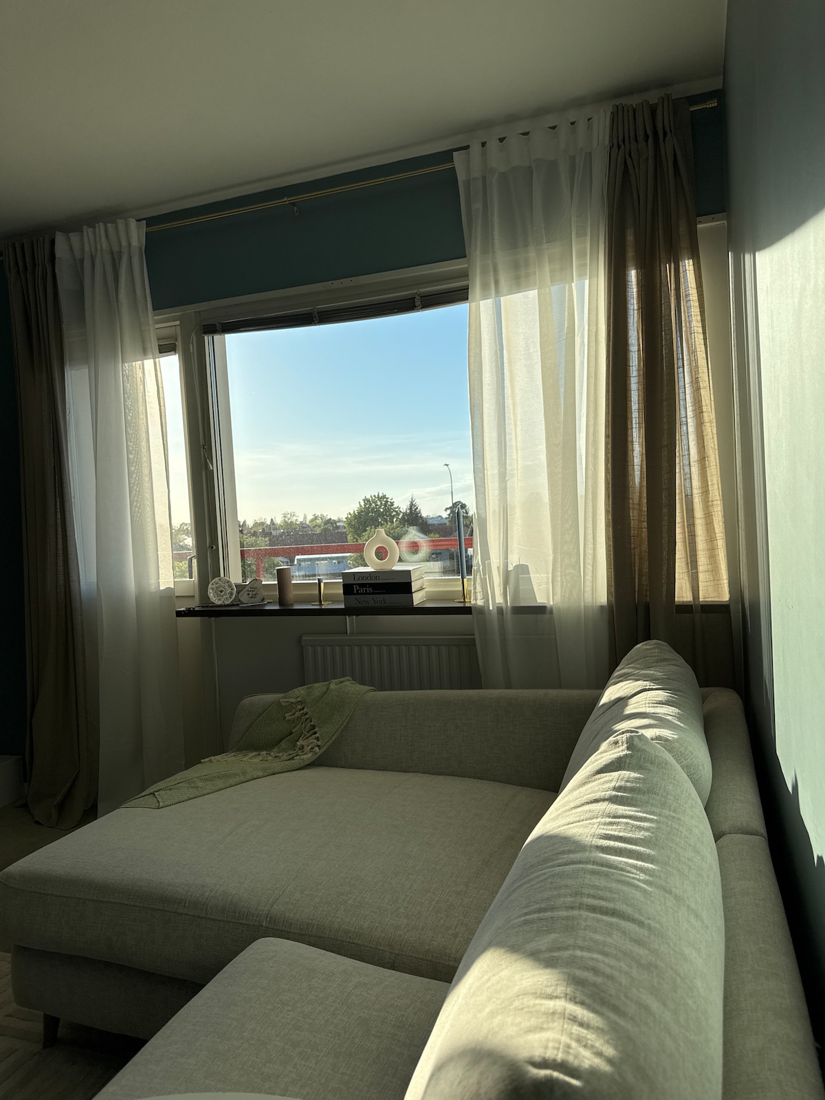 A corner of the room showcases a light-colored sectional sofa, positioned near a large window. The window reveals a view of the outdoors, framed by sheer and solid curtains that gently filter sunlight into the space.