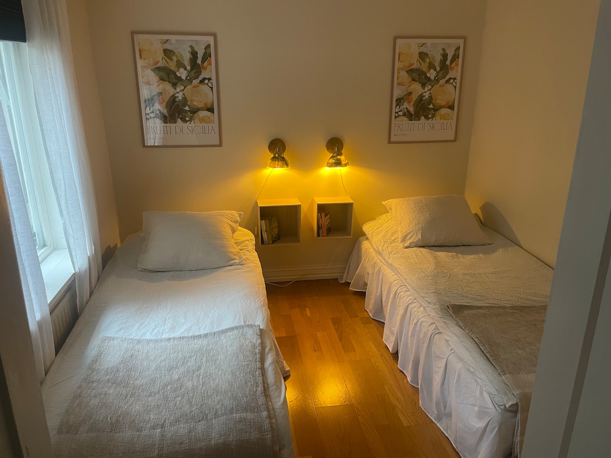 A cozy sleeping area is revealed, featuring two twin beds dressed in white linens. The warm glow from two wall-mounted lamps casts soft light on the room. Artwork is displayed above each bed, while a narrow shelf holds additional decorative elements.