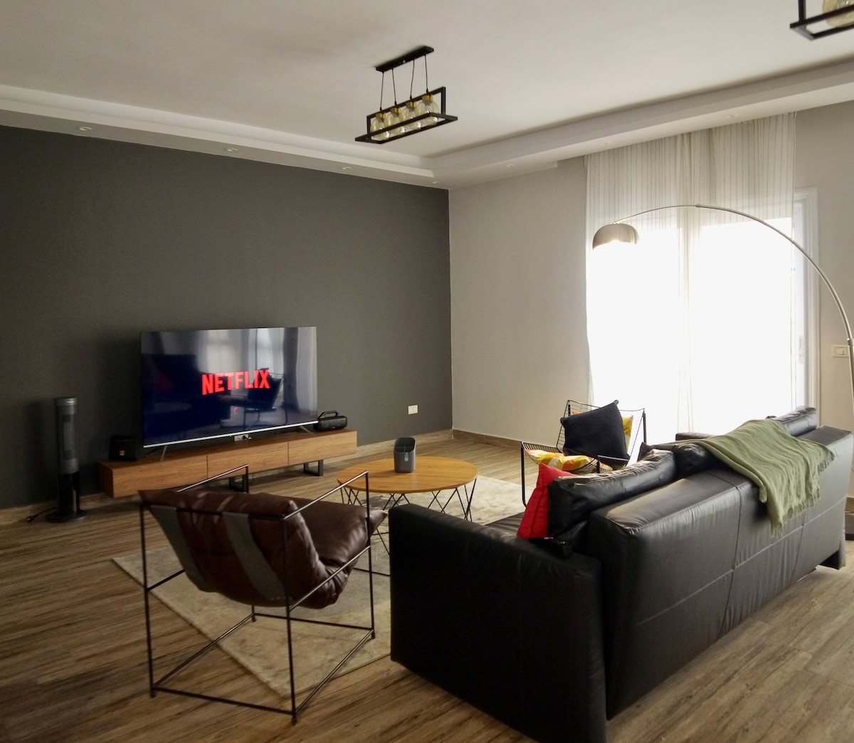 A modern living area features a large wall-mounted television displaying a streaming service. Comfortable seating, including a black sofa and wire chairs, surrounds a wooden coffee table. Natural light enters through glass doors, illuminating the room's neutral tones and wooden flooring.