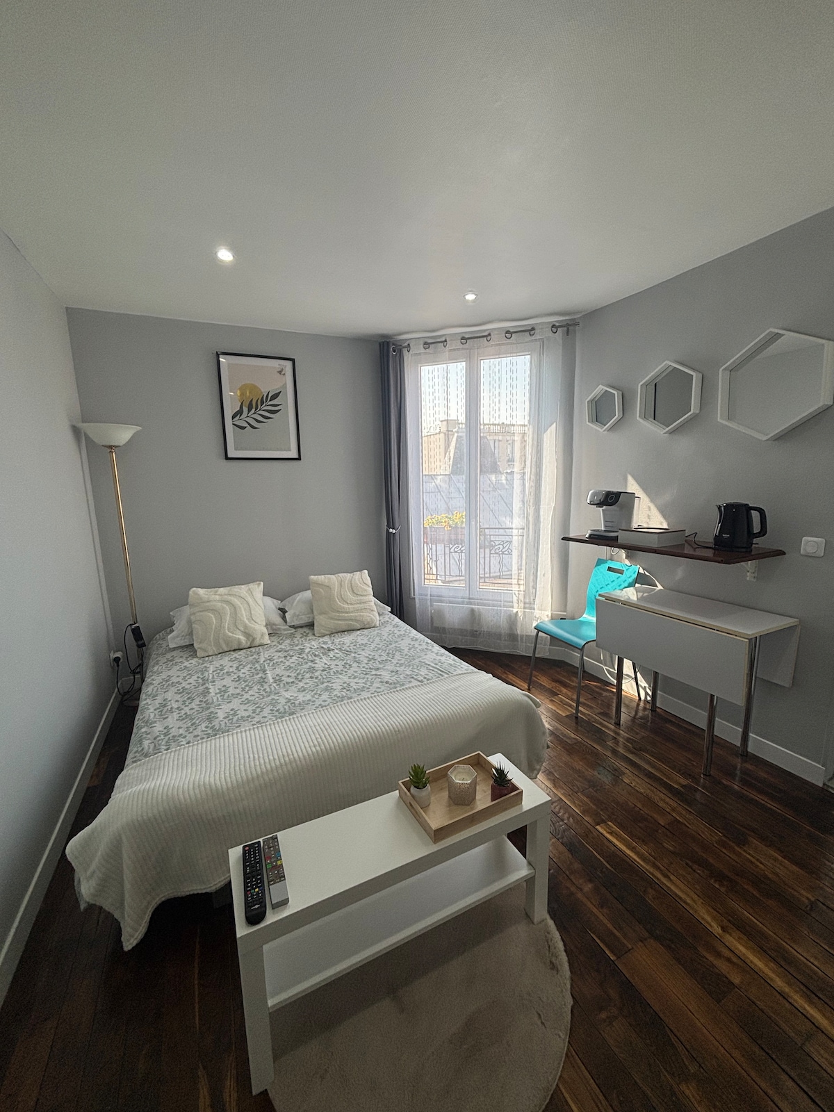 The studio features a well-lit sleeping area with a spacious bed centered against the wall. A small desk and chair are positioned beside a window, which is dressed with light curtains. Decorative wall hexagons add character, while a small coffee setup is visible to the side.