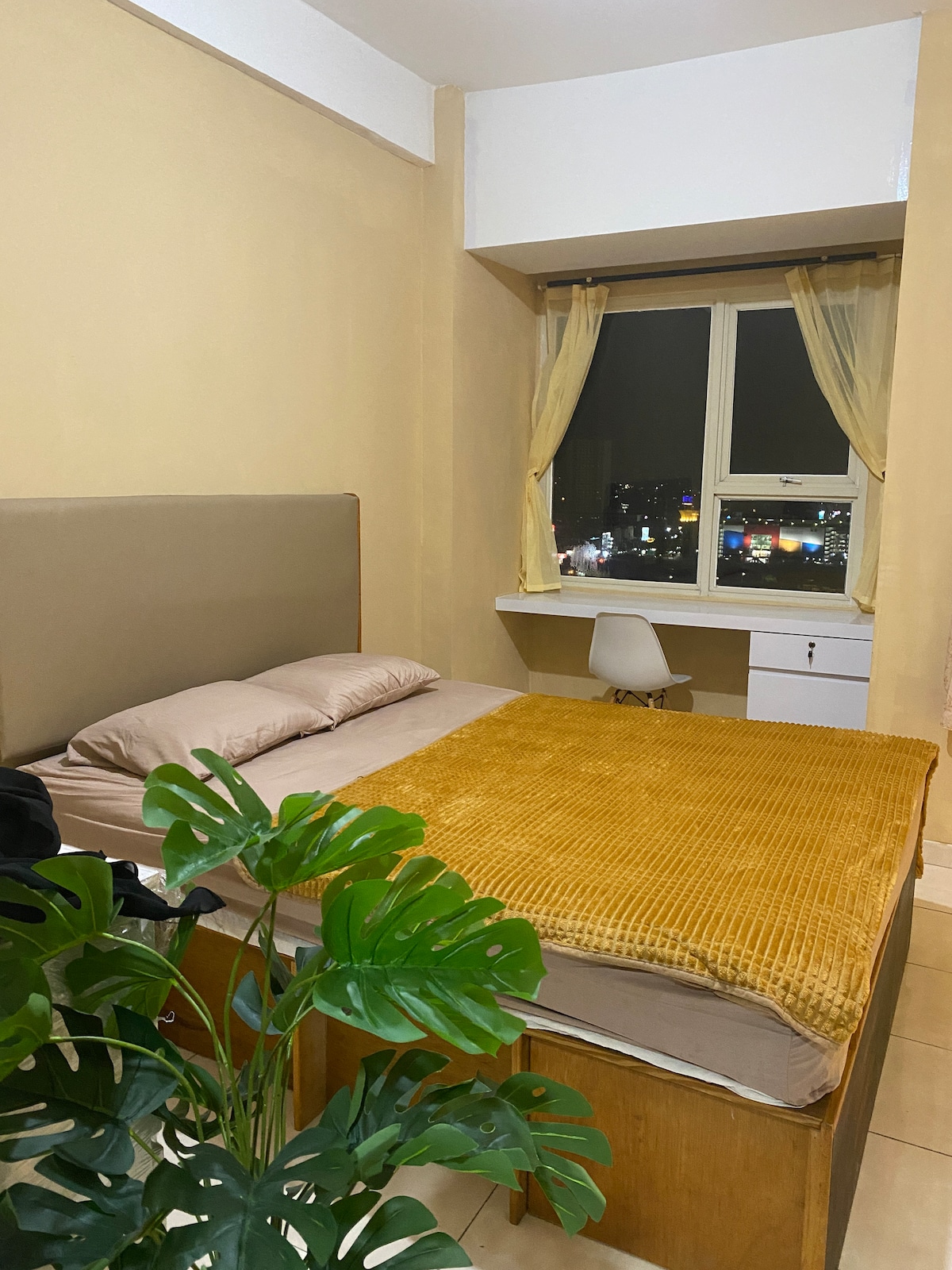A cozy bedroom features a spacious bed with a neutral-toned blanket. Soft curtains frame a large window, offering views of the city lights at night. A simple chair and a white cabinet are present, along with a green plant adding a touch of nature.