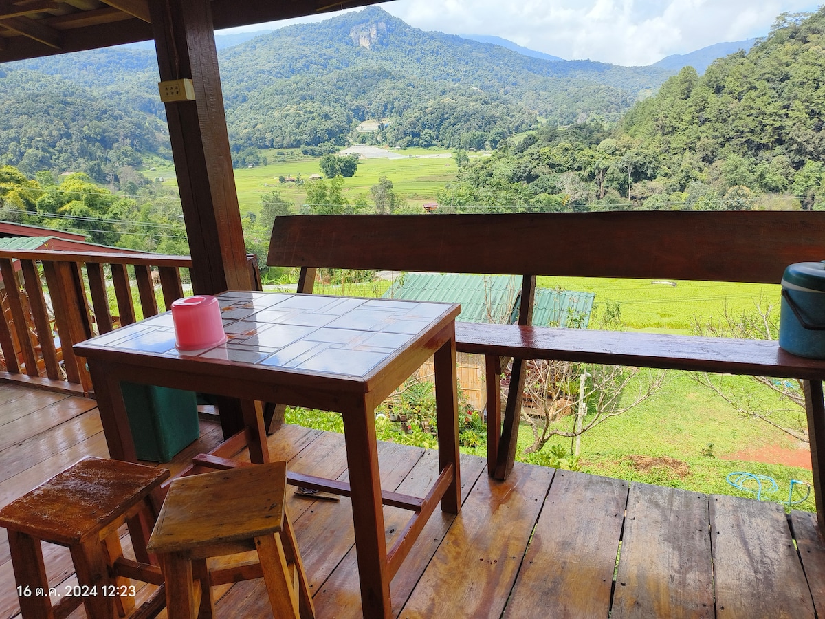 A wooden terrace is featured, offering a table with a tiled surface and benches overlooking lush green rice fields and distant mountains. Natural light floods the area, enhancing the scenery framed by wooden railings.